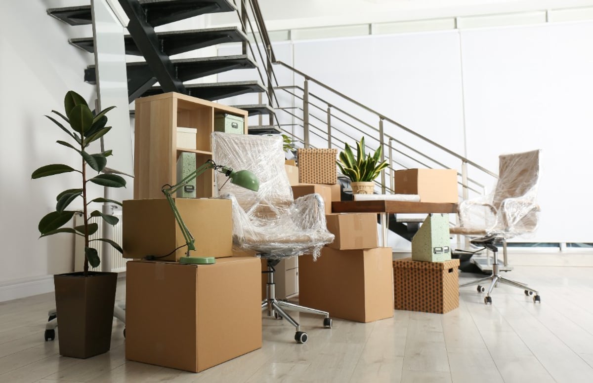 Office move day staging with cardboard boxes, plastic-wrapped chairs, plants, and a modern staircase